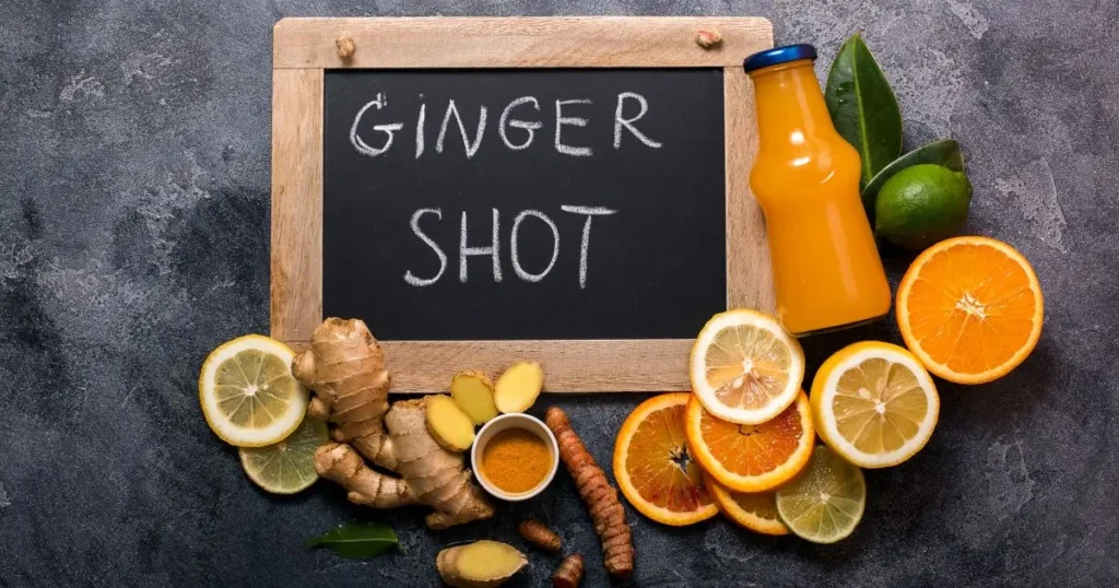 Fresh ginger shot ingredients including ginger root, citrus fruits, turmeric, and a bottle of orange juice with a chalkboard that reads 'Ginger Shot'.