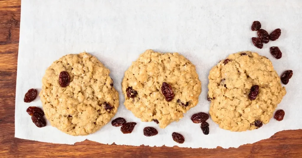 Three oatmeal raisin cookies on a parchment-lined wooden surface, surrounded by scattered raisins.